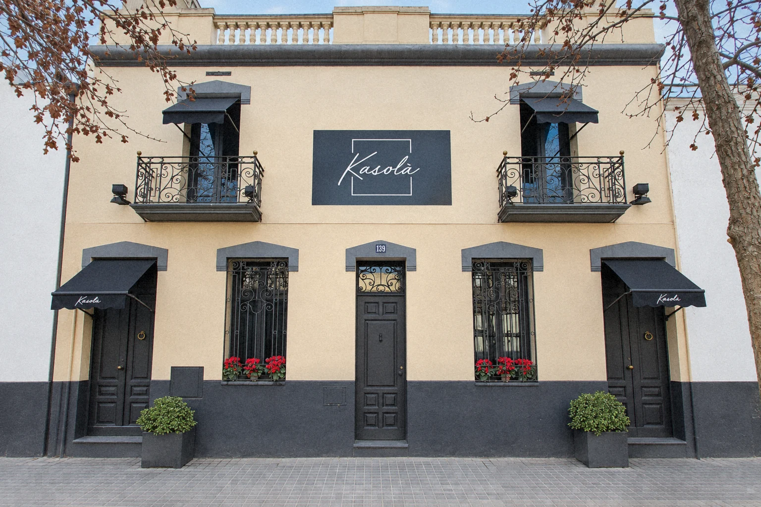 Façana de Kasolà: casa antiga de parets clares, portes negres, toldos amb el logo, balcons de ferro forjat i flors vermelles a les finestres